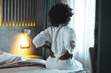 Person in a bathrobe holding their lower back while sitting on a bed.