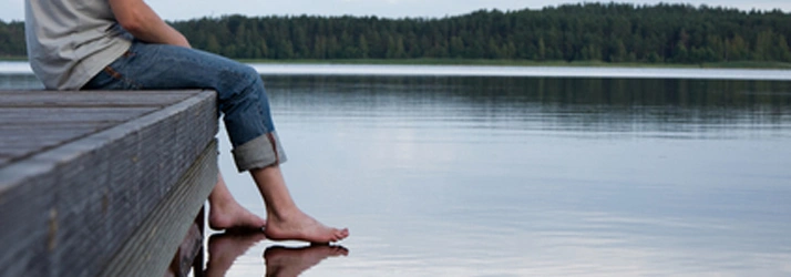 Person sitting on a dock by a lake, touching the water with their feet.